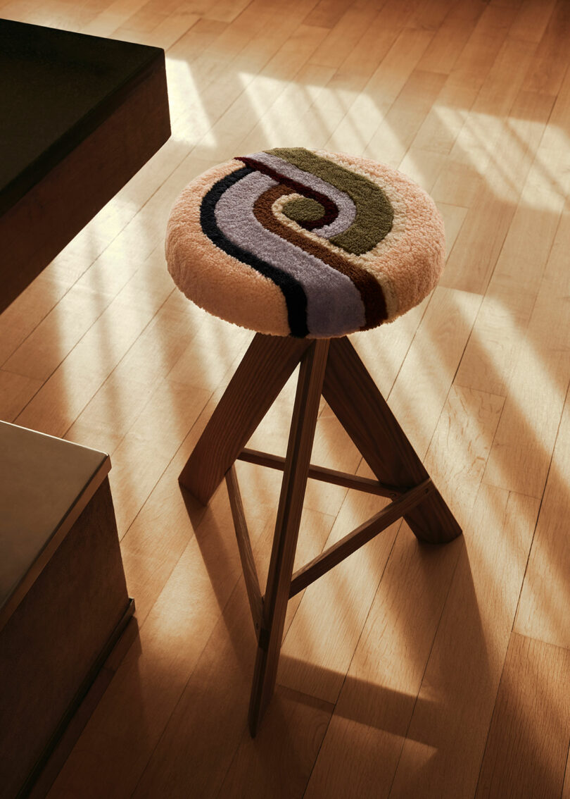 A wooden stool by Yves Salomon with a woolen, multicolored swirl-patterned cushion is positioned on a wooden floor beside a table. Sunlight casts shadows through a window onto the floor.