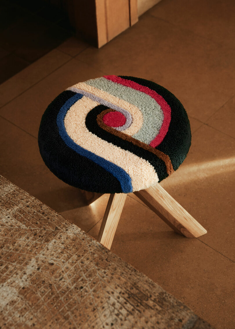 A round stool by Yves Salomon with wooden legs features a colorful, abstract patterned cushion atop it, placed on a tiled floor indoors.