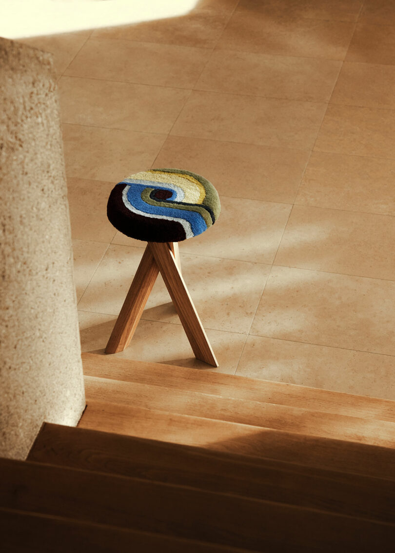 A wooden stool by Yves Salomon with a colorful round cushion is positioned near the bottom of a staircase on a tiled floor.