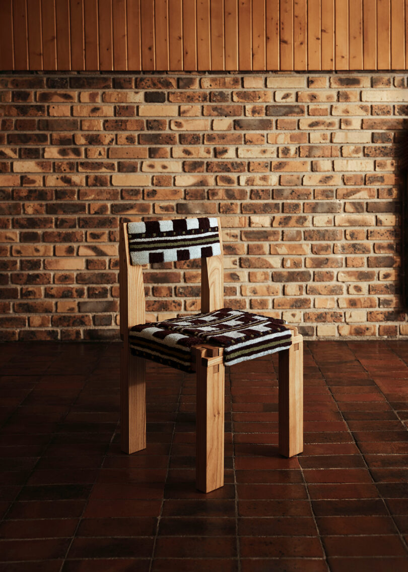 A wooden chair by Yves Salomon with a woven backrest and seat, featuring a black and white geometric pattern, placed on a tiled floor in front of a brick wall.