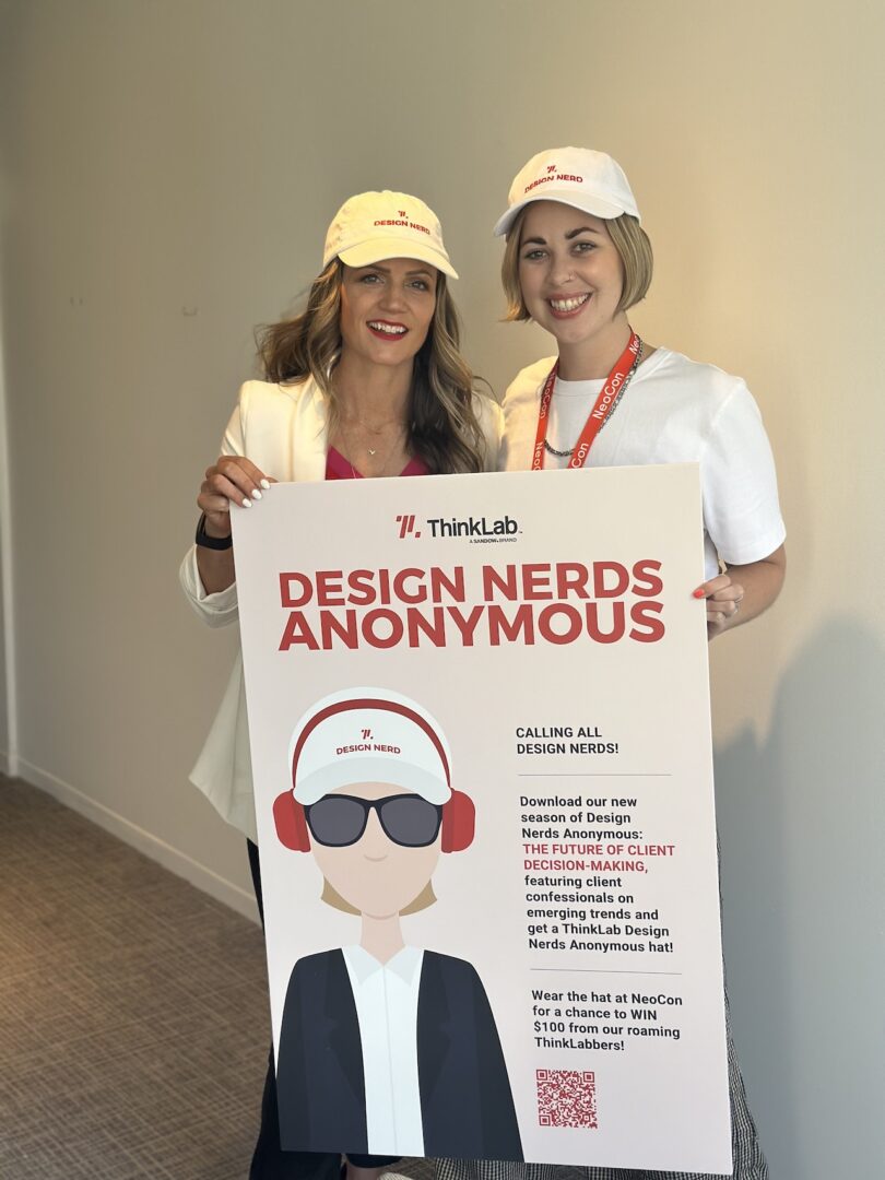 Two people wearing white ThinkLab caps holding a large poster for "Design Nerds Anonymous" with graphic illustrations and text