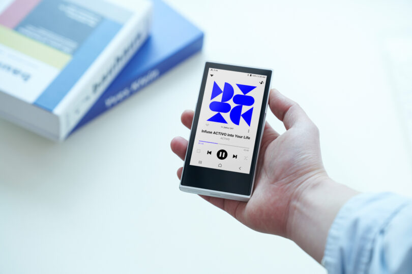 A person holds a smartphone displaying a music app with a blue geometric album cover and playback controls, while an Astell&Kern P1 sits beside a stack of books in the background on a white surface.