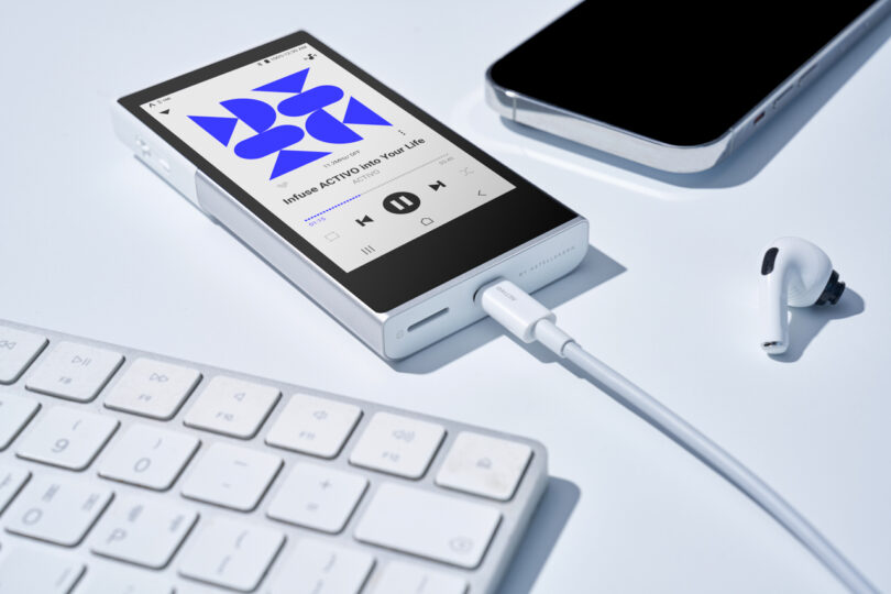 An Astell&Kern P1 portable music player, connected to a cable and playing a song, lies on a white surface near a keyboard, smartphone, and a single wireless earbud.