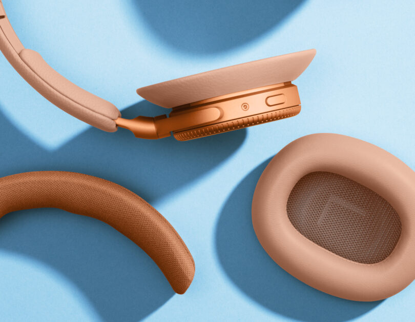 Close-up of disassembled tan-colored Beoplay H100 over-ear headphones on a light blue background, showcasing the ear cushions and headband components.