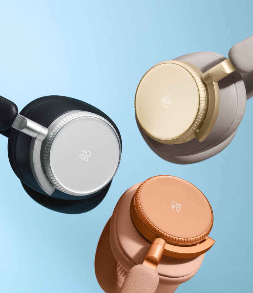 Three Bang & Olufsen Beoplay H100 headphones in black, beige, and orange colors are shown against a light blue background.