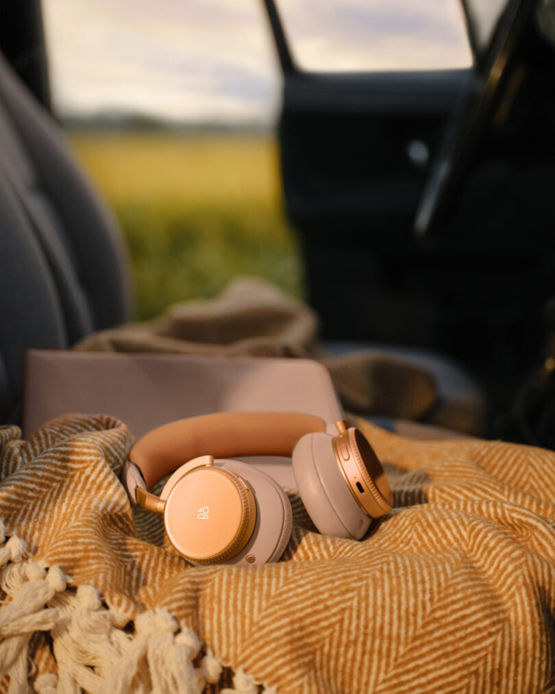 Close-up of Beoplay H100 gold and tan headphones resting on a yellow and white blanket inside a vehicle, with a blurry field visible through the window.