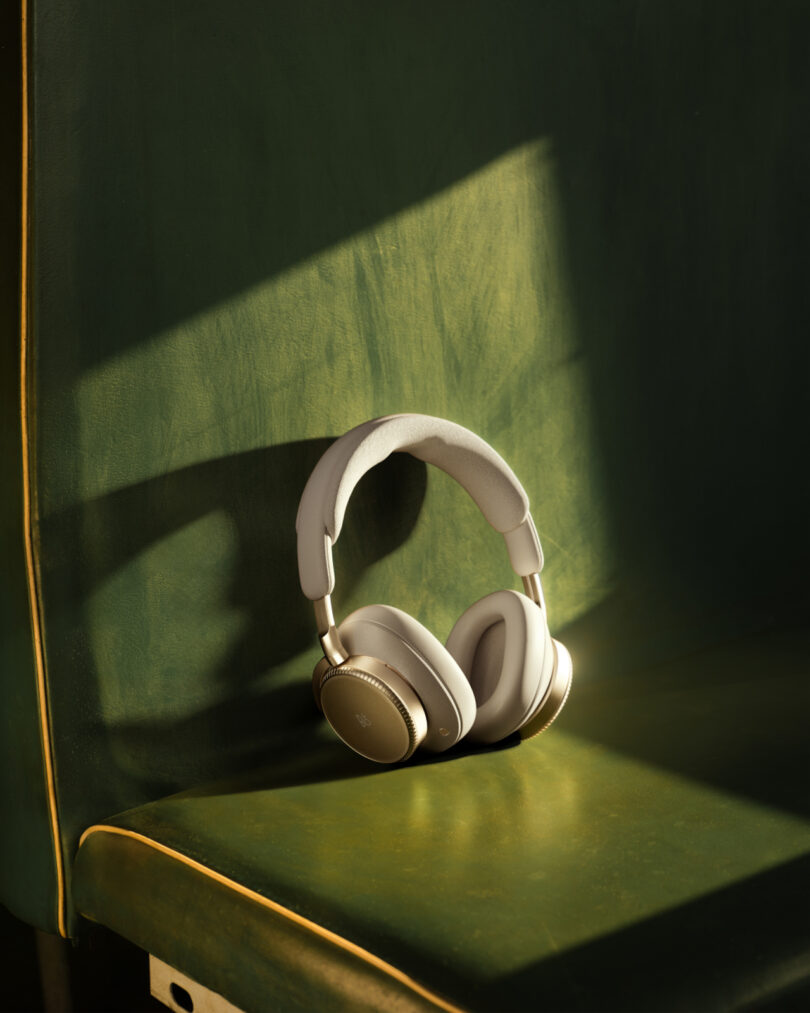 A pair of Beoplay H100 over-ear headphones rests on a green upholstered seat, partially illuminated by sunlight.