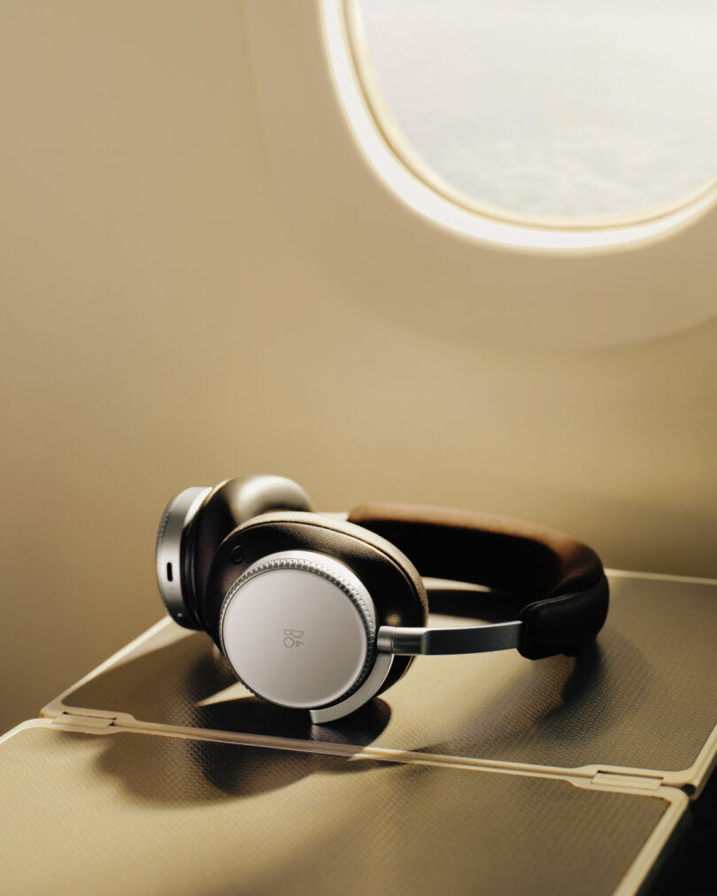 Over-ear Beoplay H100 headphones rest on an airplane tray table in front of an illuminated window.