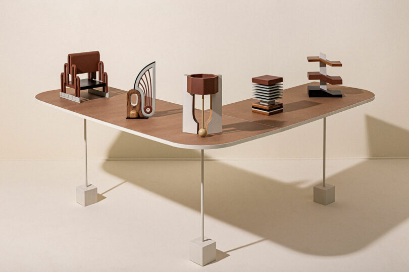 A modern table with white legs features five abstract architectural models in various shapes and colors, primarily in brown and neutral tones, arranged on a matte wooden surface