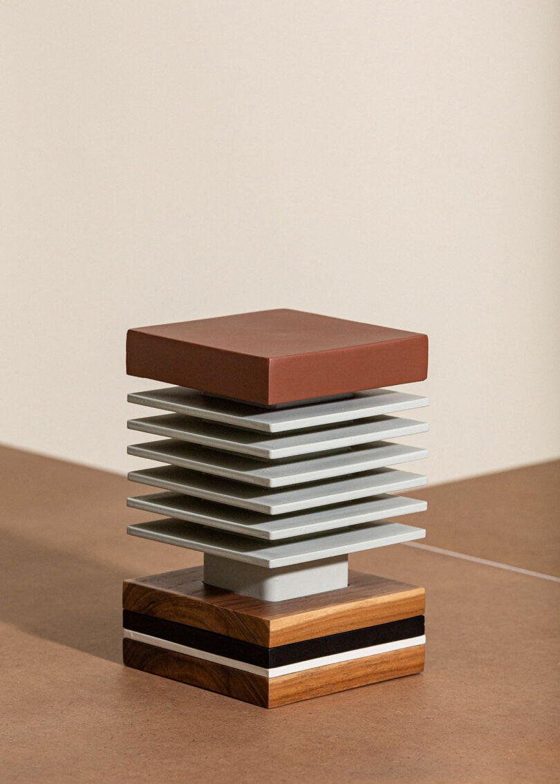 A geometric sculpture with alternating beige and brown layers, featuring a wooden base with black, white, and wood stripes