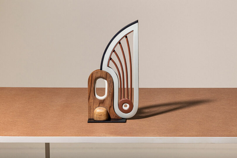 A modern abstract sculpture featuring geometric shapes in wood, metal, and plastic sits on a flat, brown surface against a neutral background