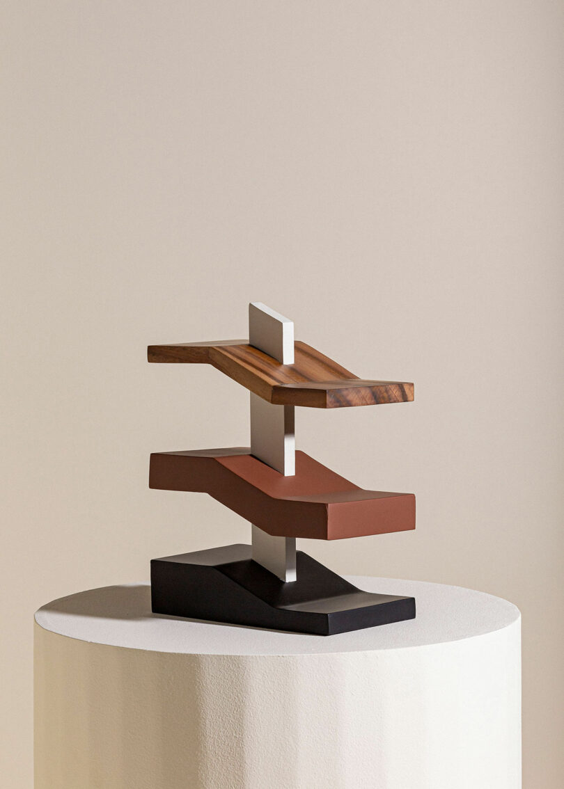 A minimalist, stacked modern sculpture with interlocking rectangular blocks in different colors and materials, displayed on a round pedestal against a neutral background