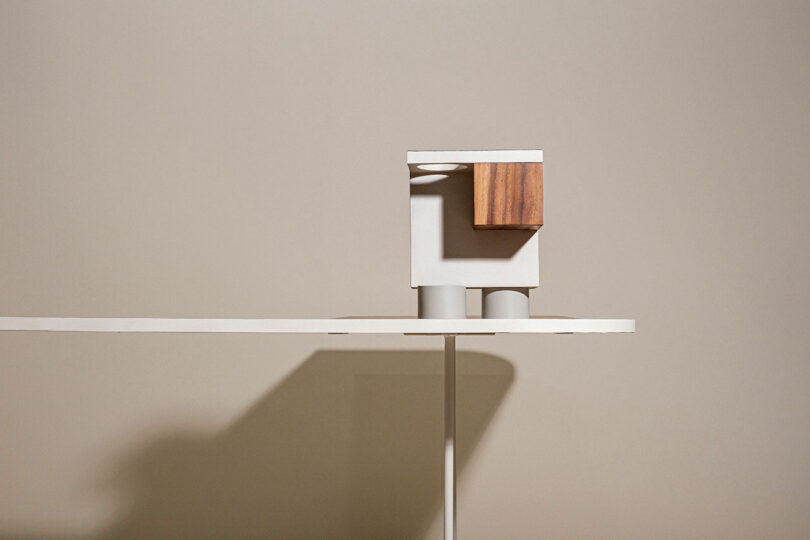 A modern minimalist sculpture featuring abstract geometric shapes, including a white block, a wooden piece, and cylindrical elements, displayed against a beige wall on a white shelf