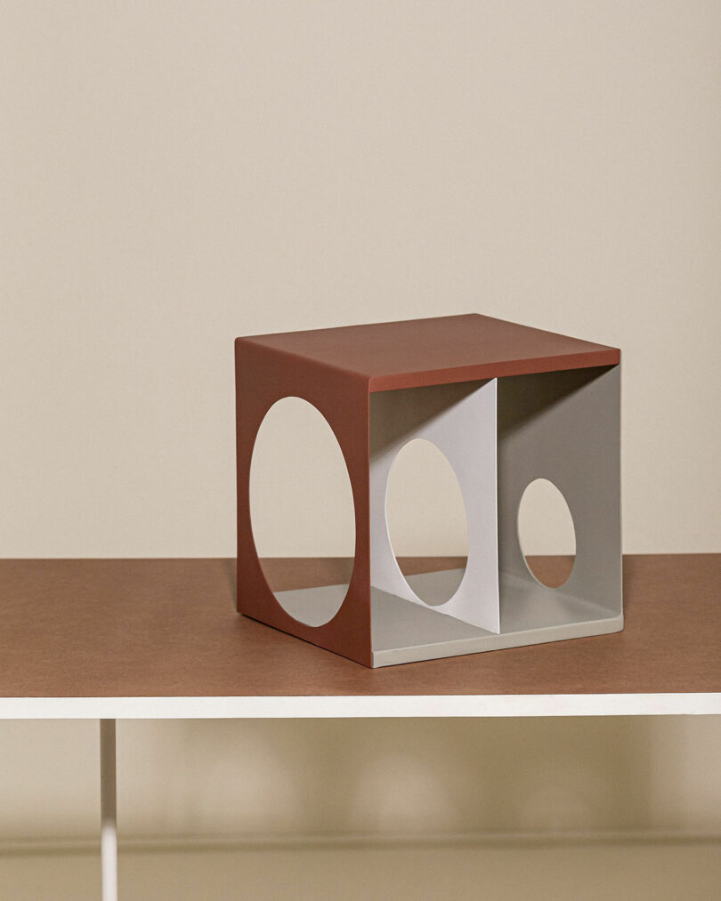 A geometric sculpture featuring a rectangular form with circular cutouts on its side rests on a wooden surface against a beige backdrop