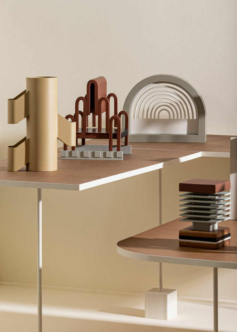 A modern art display featuring abstract geometric sculptures in brown, beige, and gray tones, arranged on white and wooden surfaces