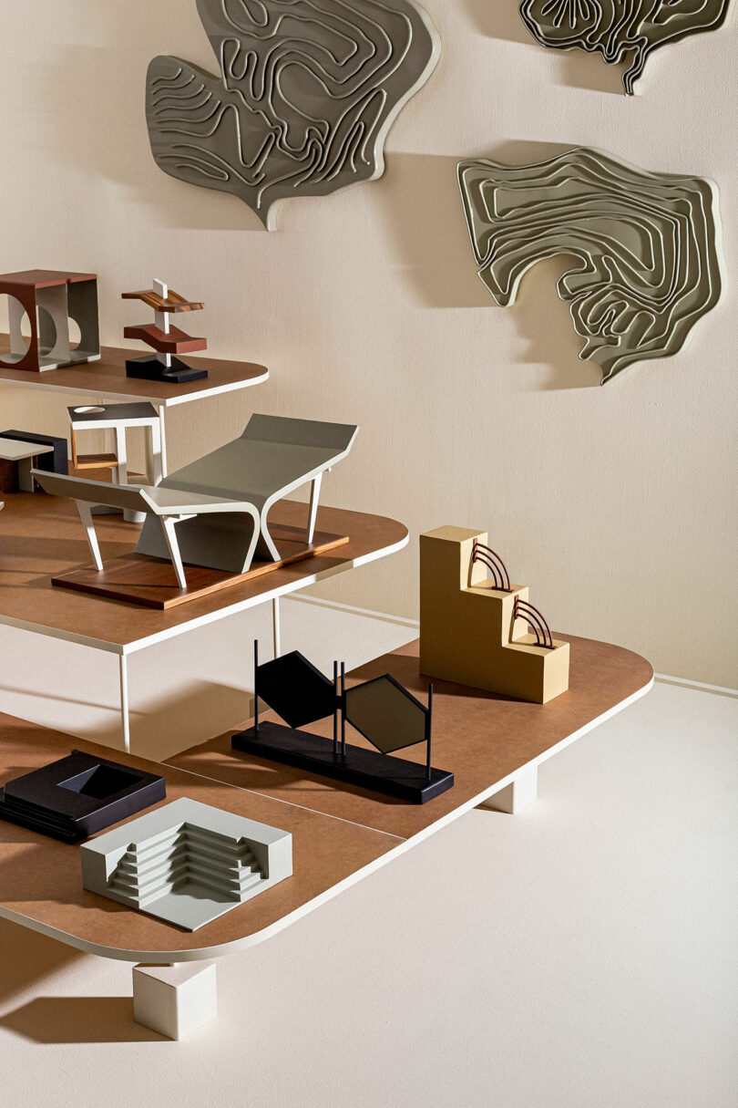 A geometric display of abstract architectural models on tiered wooden shelves, set against a wall decorated with sculpted, organic shapes