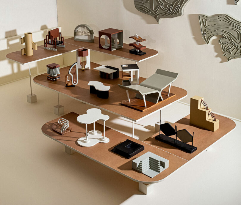 A display of abstract architectural models arranged on three wooden shelves against a beige background