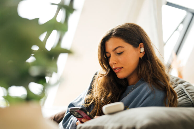 A woman with long hair, wearing earbuds, looks at her smartphone while sitting on a sofa. A plant is partially visible in the foreground.