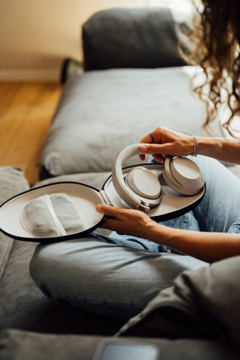 Person sitting on a couch, holding a white over-ear headphone set inside a white carrying case. The person's face is not visible.