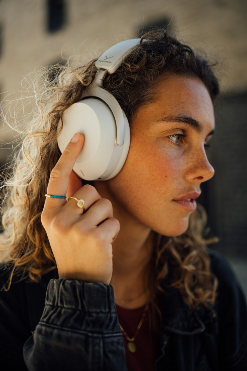 A woman with curly hair wearing over-ear headphones and touching the left earpad, standing outdoors.