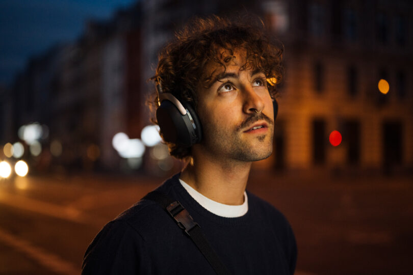 Person with curly hair wearing headphones and a shoulder strap looks upward on an urban street at dusk.