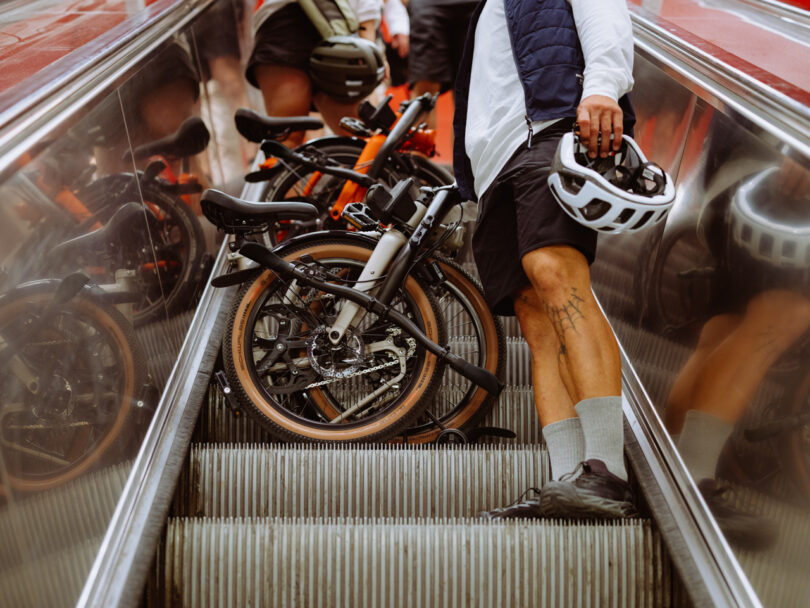 Two people holding folded bicycles stand on an escalator. One person holds a helmet. Their legs and bicycles are the main focus. The background consists of reflective escalator walls and handrails.