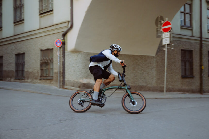 A cyclist wearing a helmet and backpack rides a bicycle on a city street near a building with barred windows. A no-entry road sign is visible in the background.