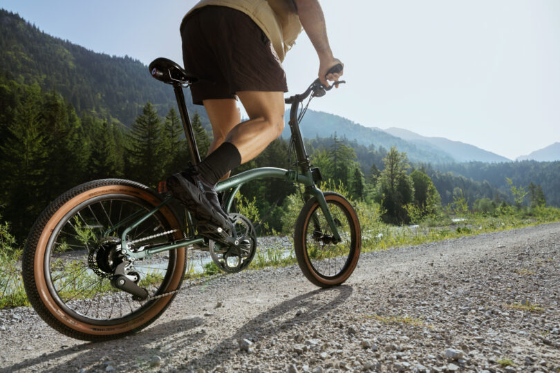 Person riding a green bicycle on a gravel path in a mountainous forest area under a clear sky.