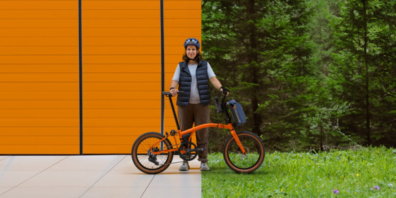 A woman stands next to an orange bicycle, with a background split between an orange wall on the left and a forest on the right.