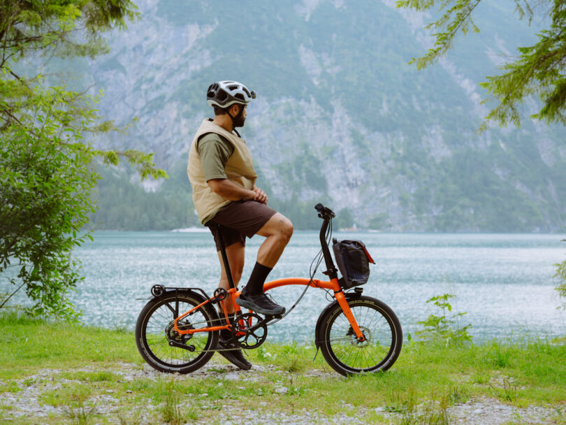 A person wearing a helmet and vest sits on an orange bicycle, overlooking a serene, mountainous lake surrounded by greenery.