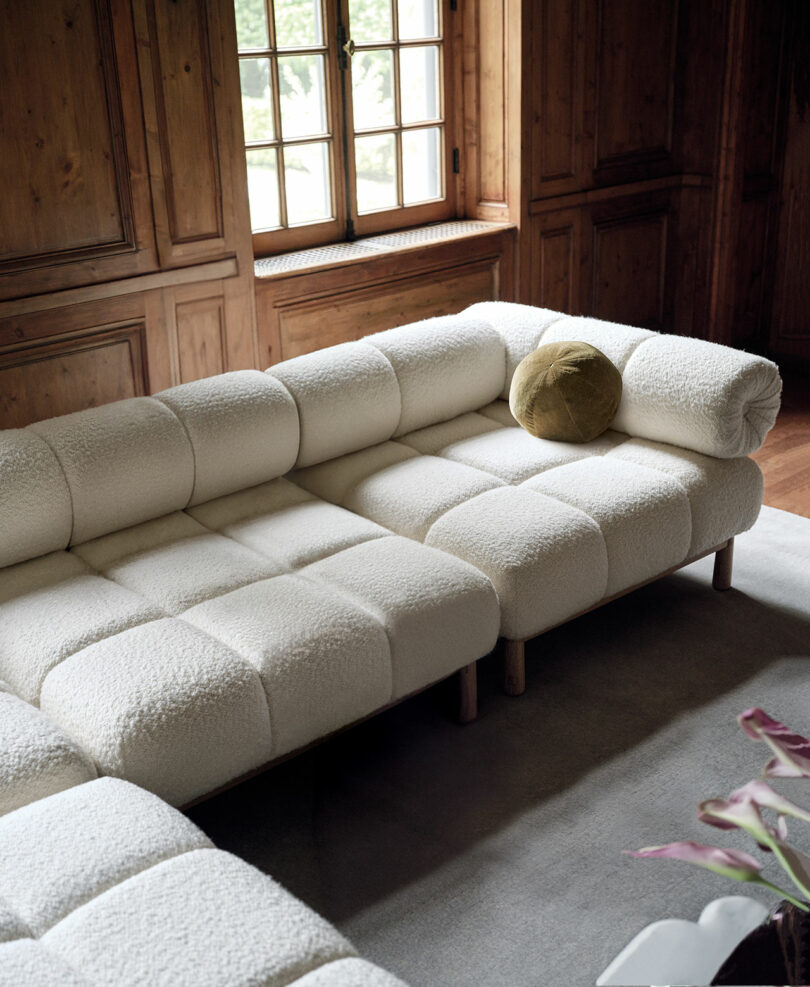 A modern beige sectional sofa with tufted cushions sits in a cozy wooden-paneled room. A green round pillow rests on the sofa