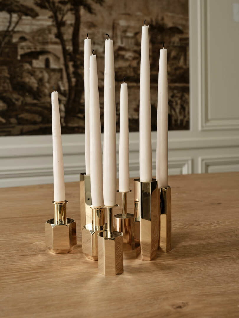 A group of tall, white taper candles are held in modern, reflective gold candle holders set on a light wooden table. In the background is a decorative wall tapestry