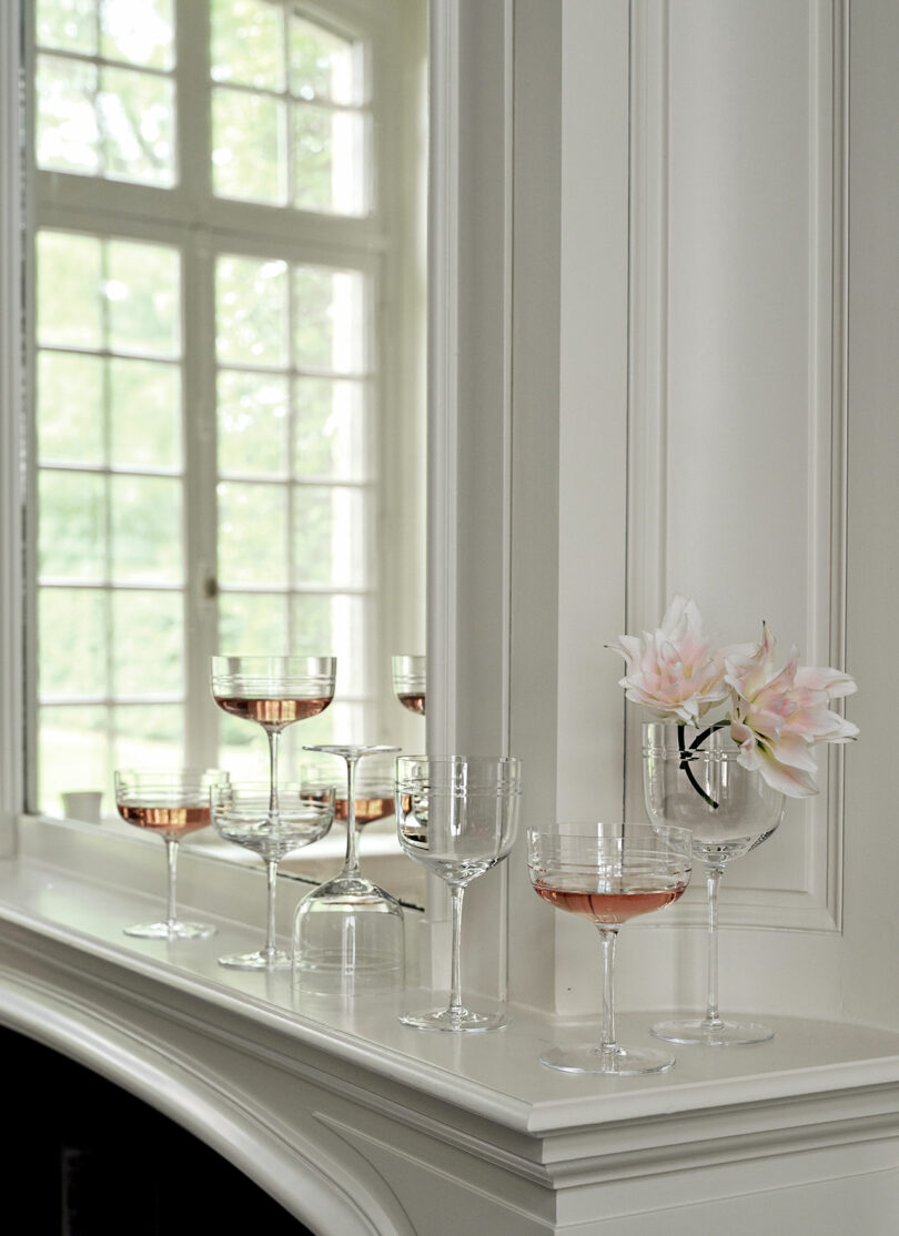 A collection of wine glasses, partially filled with pink liquid, is arranged on a white mantel. A large window and pale pink flowers in one glass are also visible in the background