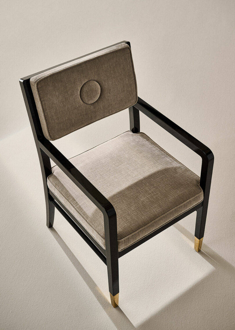 A beige cushioned chair with a dark wooden frame and brass-tipped legs is placed on a light-colored floor