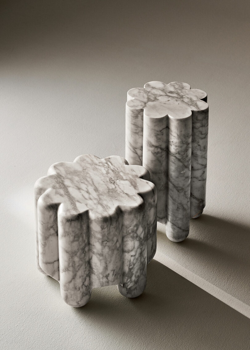 Two sculptural marble stools with flower petal designs sit on a smooth surface in a neutral tone. The light casts soft shadows, highlighting the intricate details of the stone