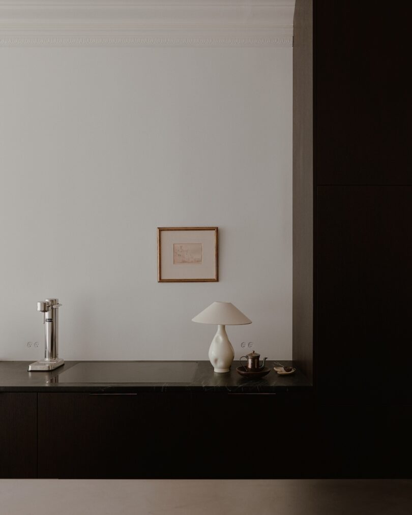 Minimalist kitchen with dark cabinetry, a white wall, and a black countertop. A small framed picture, a white table lamp, and a few kitchen items are seen on the counter.