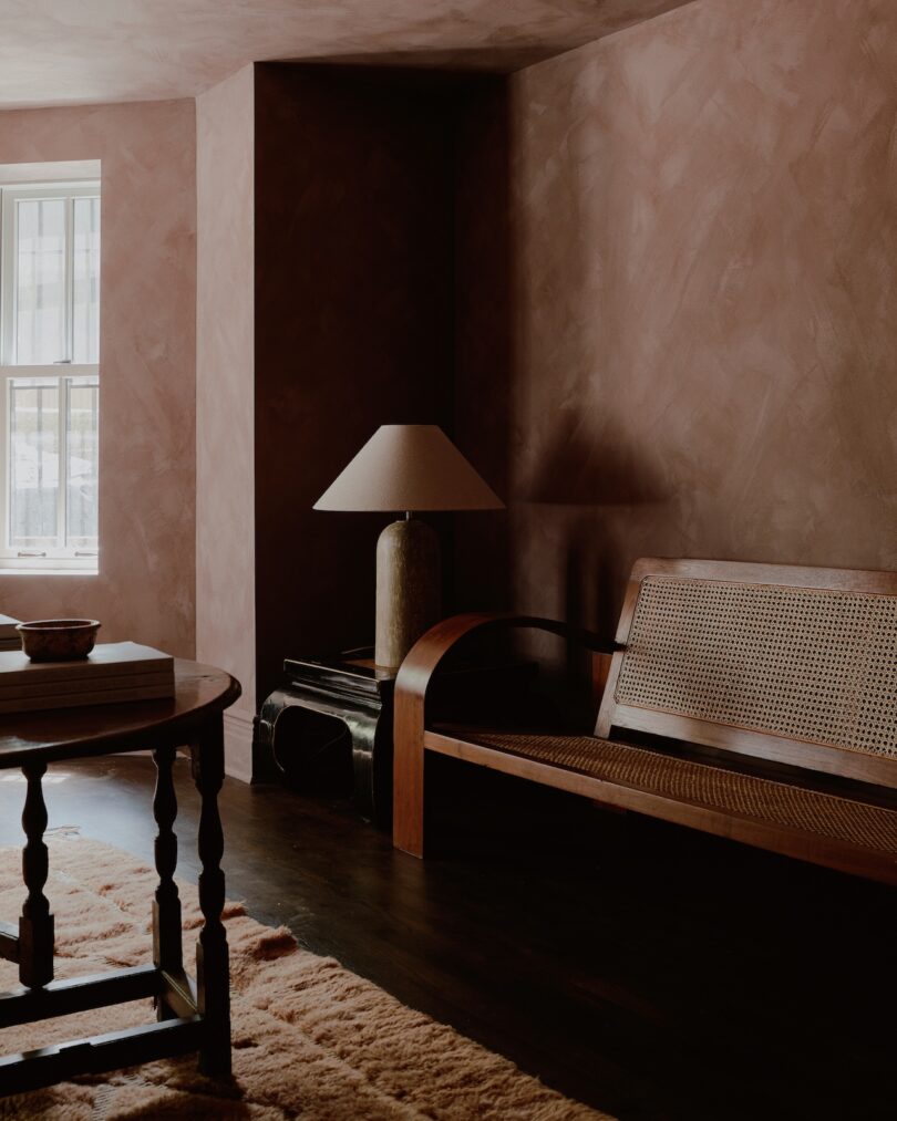 A dimly lit room with brown walls features a table, a woven bench, a lamp, and a window with light filtering through.