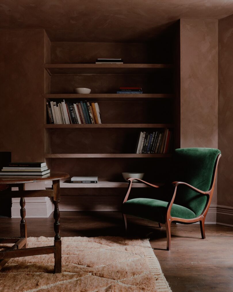 A green armchair sits in front of a built-in bookshelf filled with books and a few decorative items. A wooden table with stacked books is on the left, and a light brown rug covers the floor.