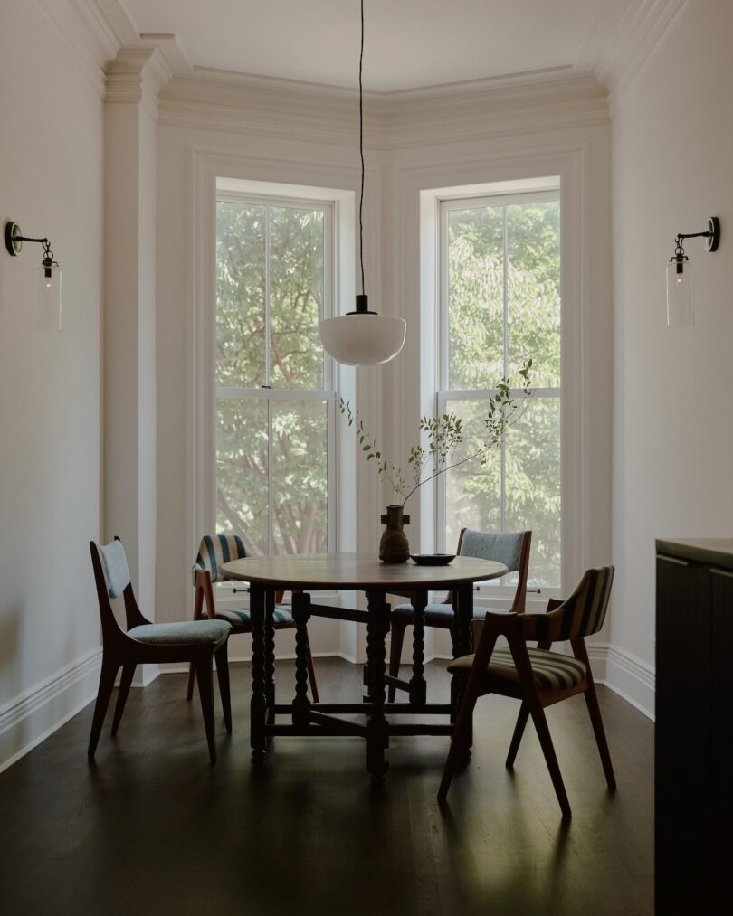 A dining area features a round table with four mid-century modern chairs, a central pendant light, and two large windows with a view of greenery outside. A vase with leafy branches is on the table.