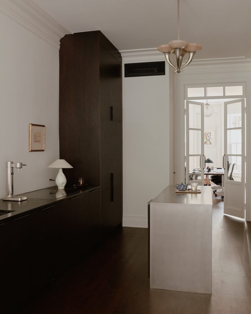 Modern kitchen with dark cabinets, a small sink, a white lamp, and stainless steel island. A door leads to a bright room with a desk and an office chair in the background.