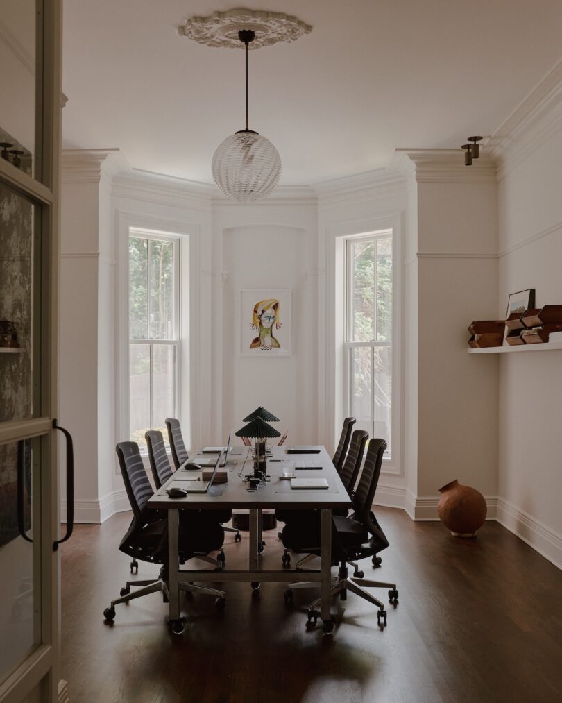 A modern conference room with a long table, black chairs, a round light fixture, three tall windows, a small shelf with decor items, and a portrait-style painting on the wall.