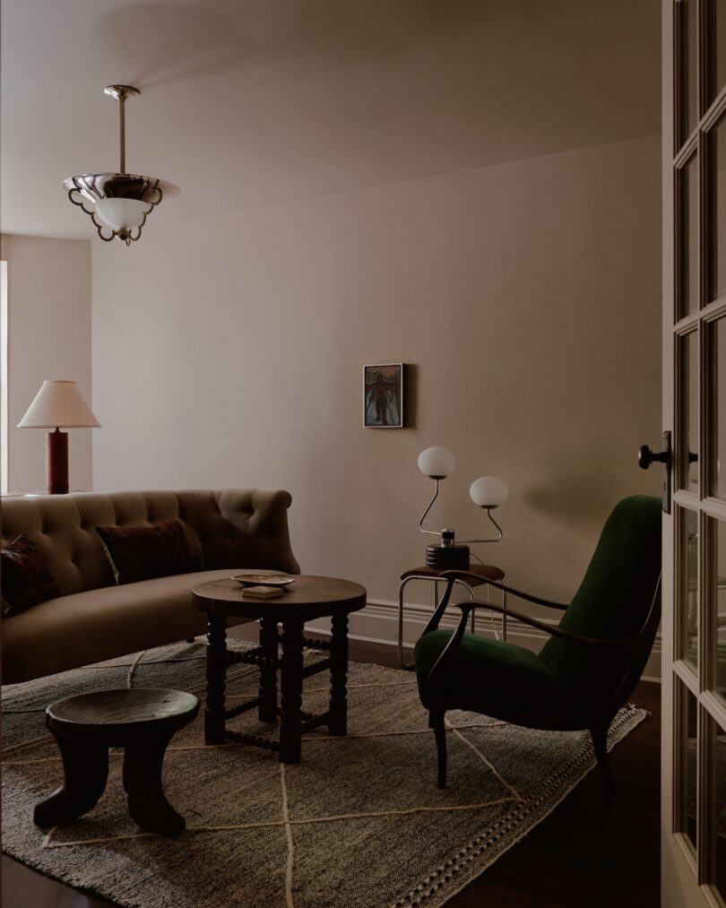 A cozy living room with a green armchair, a grey sofa, a wooden coffee table, and a small stool on a patterned rug. The room features a floor lamp, two spherical table lamps, and a wall-mounted artwork.