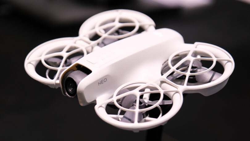 Close-up of a white quadcopter drone with propeller guards and a camera on the front. The word "NEO" is visible on the side.