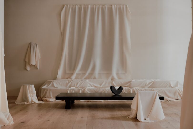 A minimalist room with white draped curtains and fabric on furniture. A black bench with a black abstract sculpture sits in front of the draped fabric. Light wooden flooring is visible.