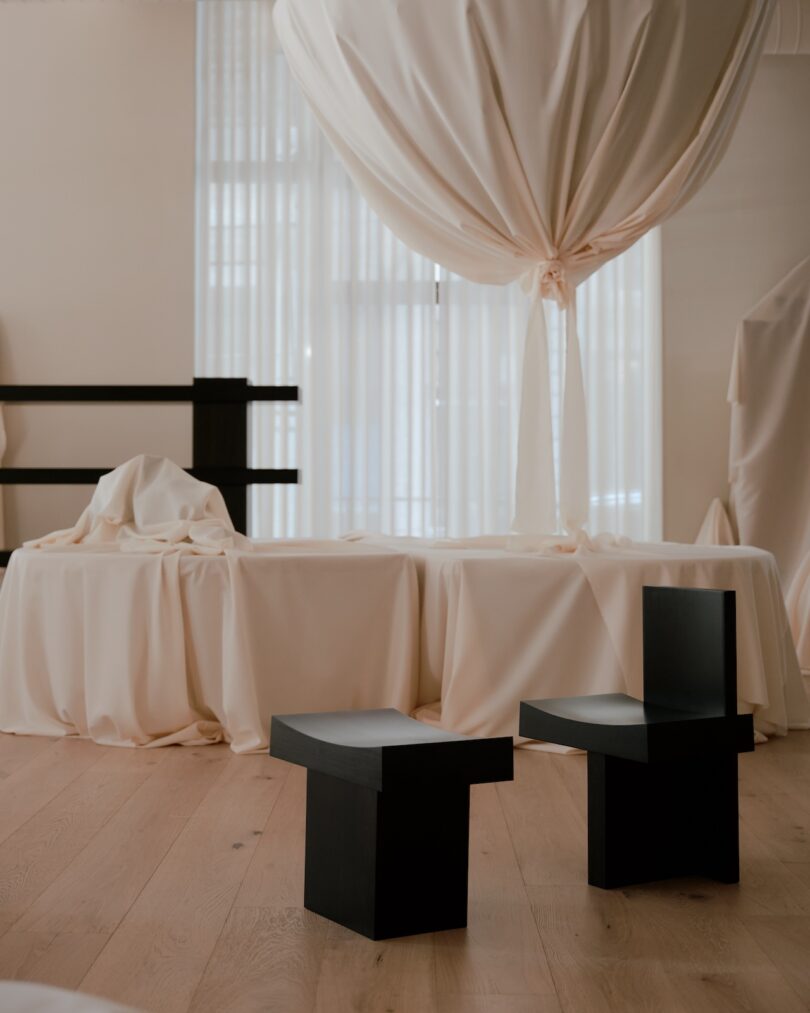 A minimalist interior with a draped table, two black geometric chairs, a cat tree, and sheer curtains.