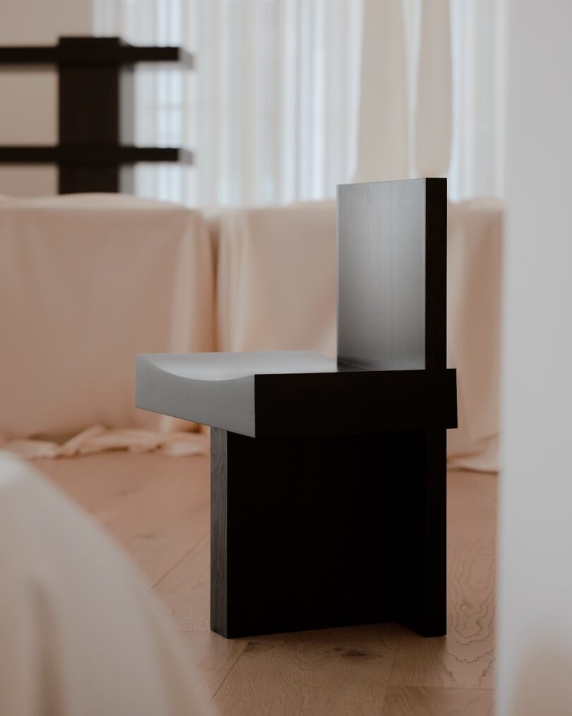 A minimalist black wooden chair is placed on a light-colored wooden floor in front of a backdrop of white curtains.