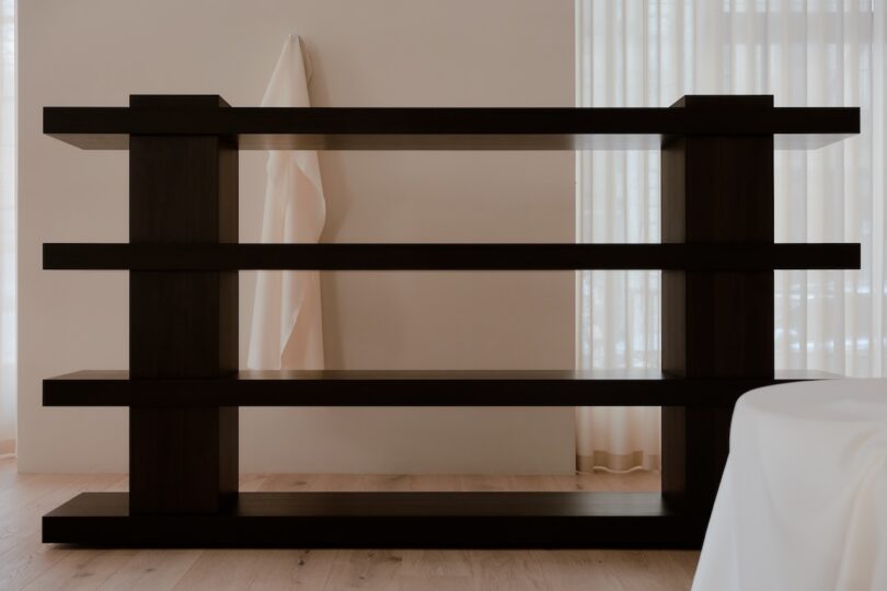 A dark wooden shelf stands against a light wall with a white cloth draped over its top corner. Light filters through sheer curtains in the background.