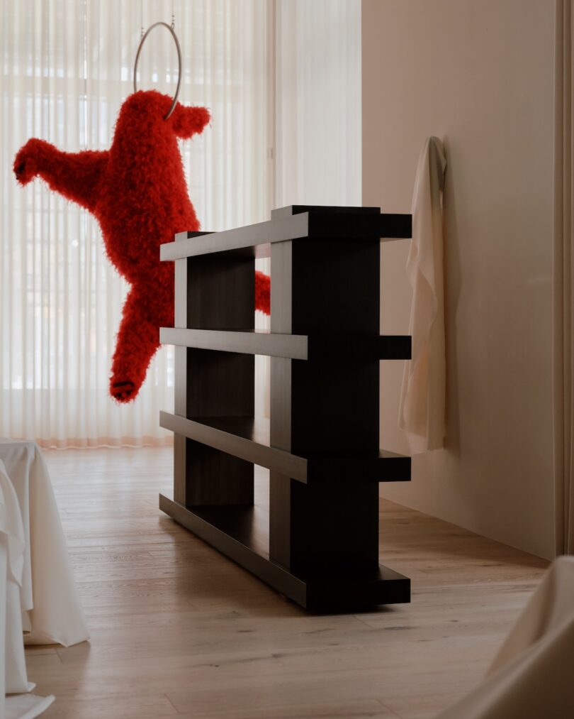 A modern dark wood shelf with several tiers stands in a minimalistic room. A red, furry figure hangs on a ring from the ceiling behind it near a large window with sheer curtains.