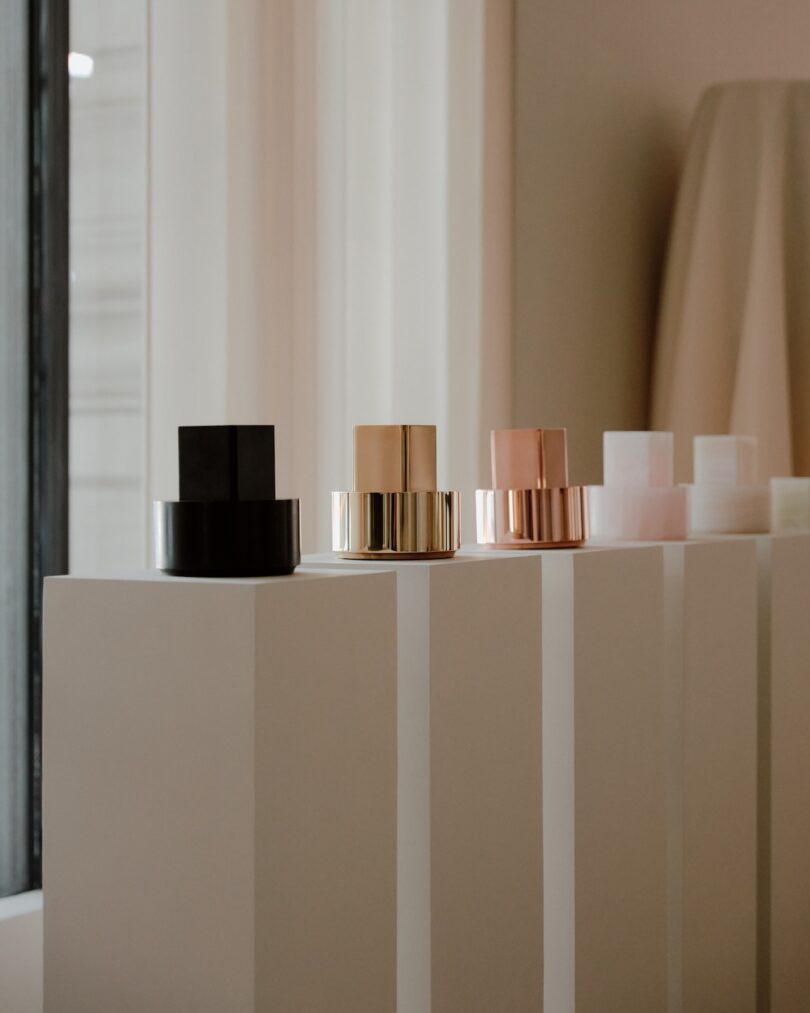 Three geometric sculptures in black, copper, and rose gold displayed on white pedestals in a minimalist room.