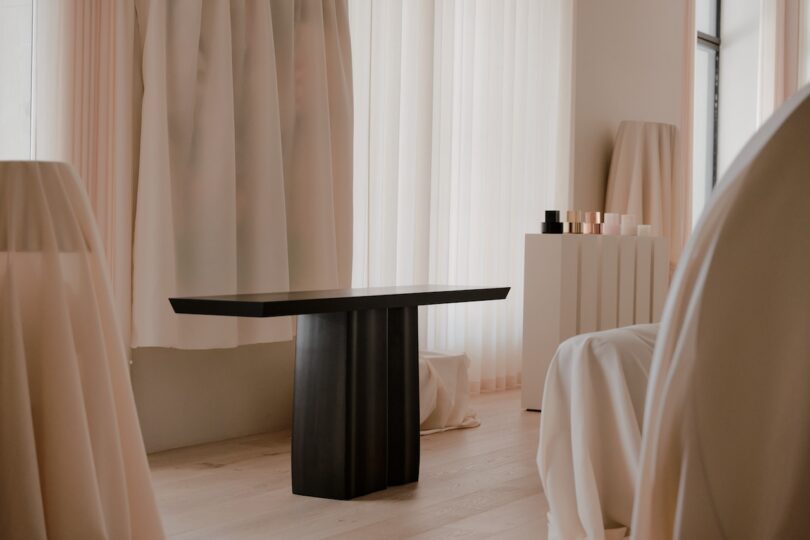 A minimalist room with a black coffee table, light wood flooring, sheer white curtains, and draped furniture in neutral tones.